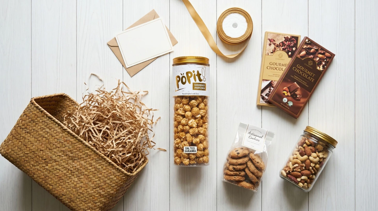 A deconstructed hamper flat-lay showing all individual items laid out before assembly. POPIT! jar included. Clean white or warm wood surface. Items arranged with breathing room.